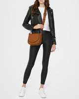A woman wearing a black leather jacket, white shirt, leggings and white shoes showing this saddle bag as a leather crossbody bag.