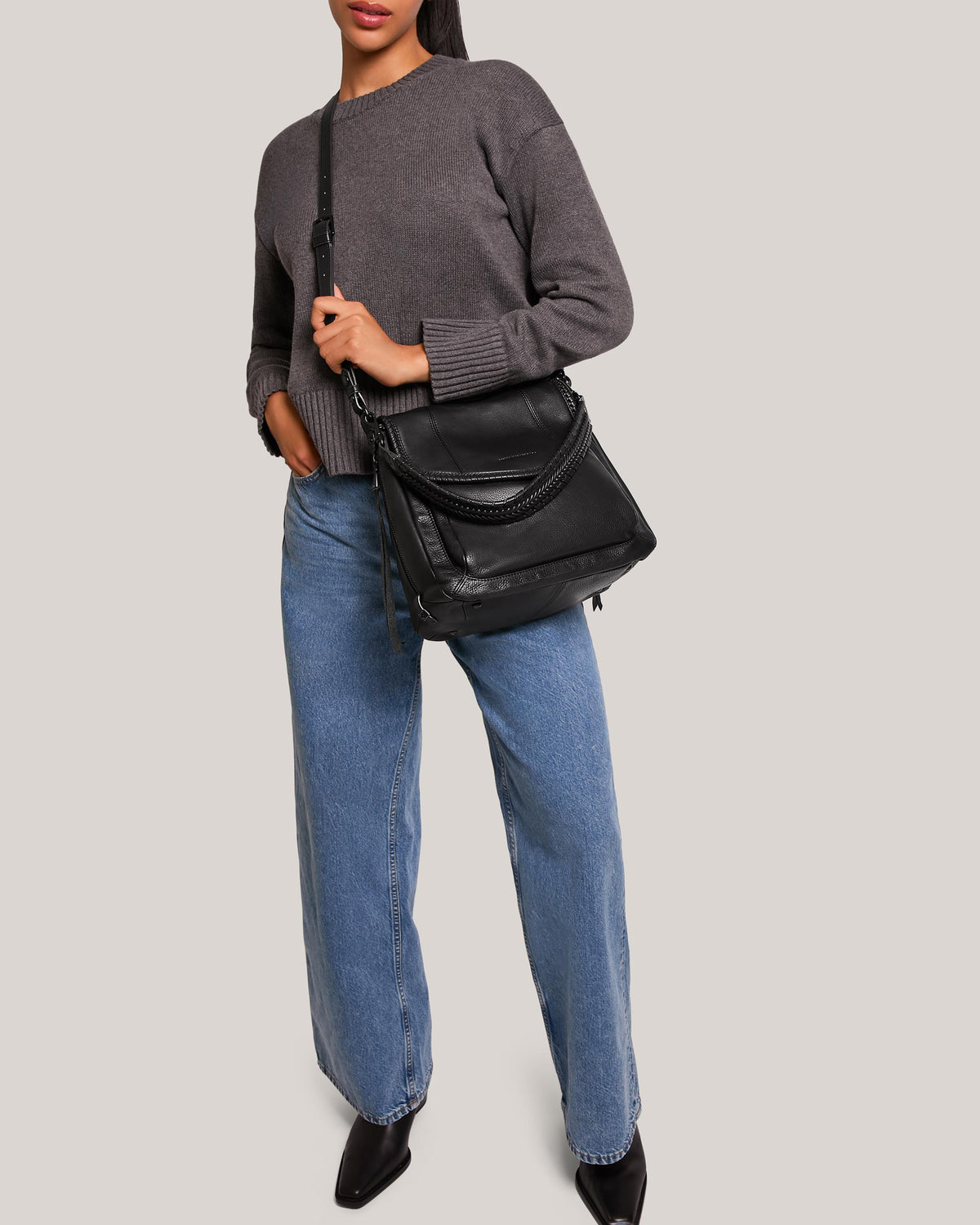 A woman in a gray sweater and blue jeans stands against a plain background, showcasing a sleek leather shoulder bag with woven leather straps