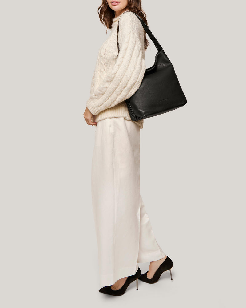A woman wearing a white sweater and pants showing off this black shoulder bag that doubles as a leather purse.