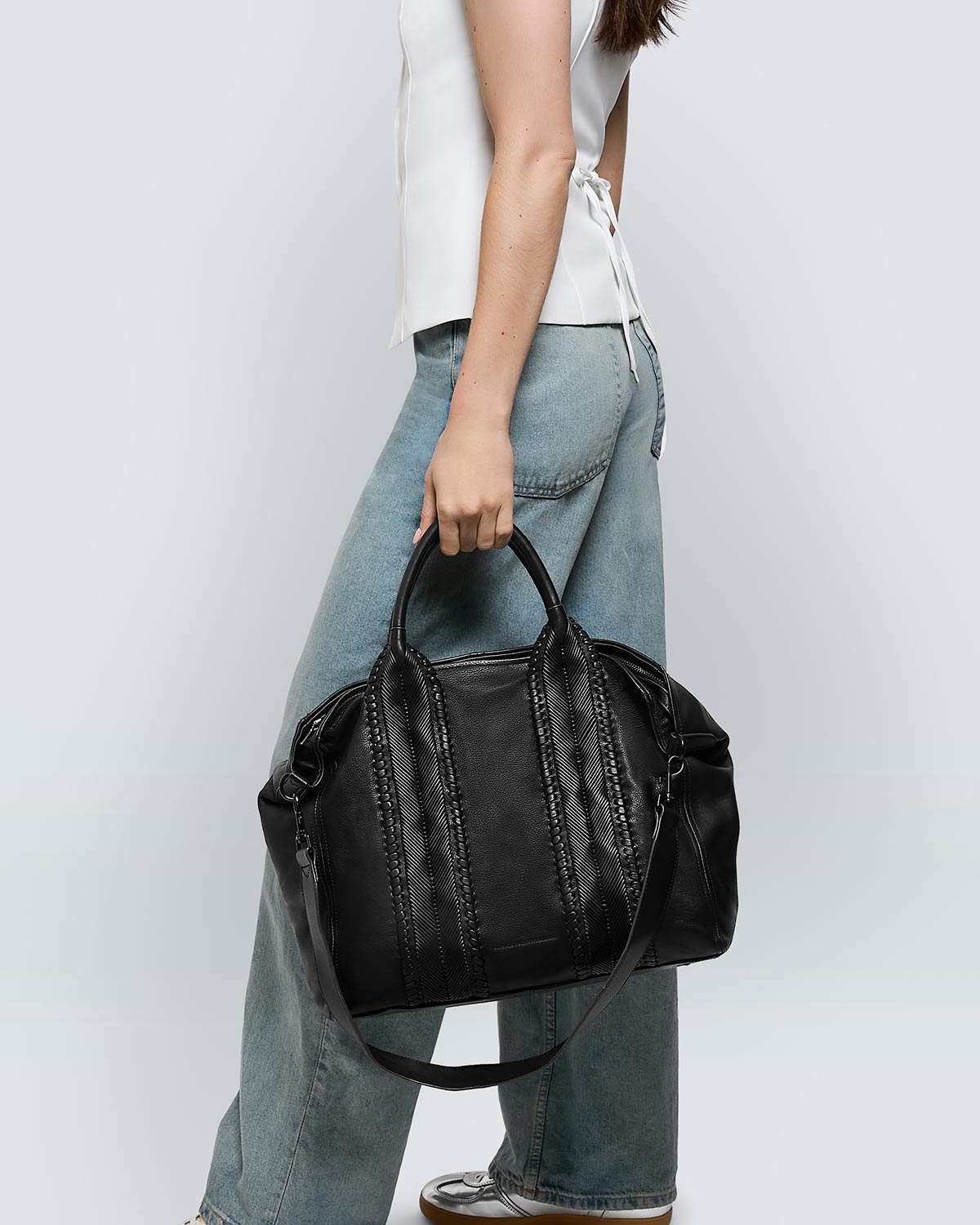 A woman wearing a white top and jeans, holding a large leather tote bag with a zipper. 