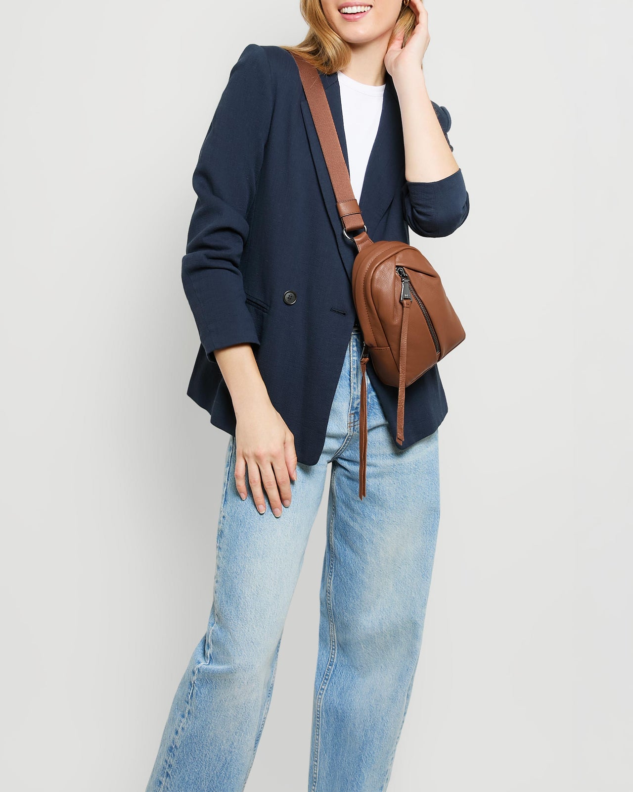 A woman wearing a blue blazer, white shirt and jeans, showcasing a leather sling bag across her chest. 